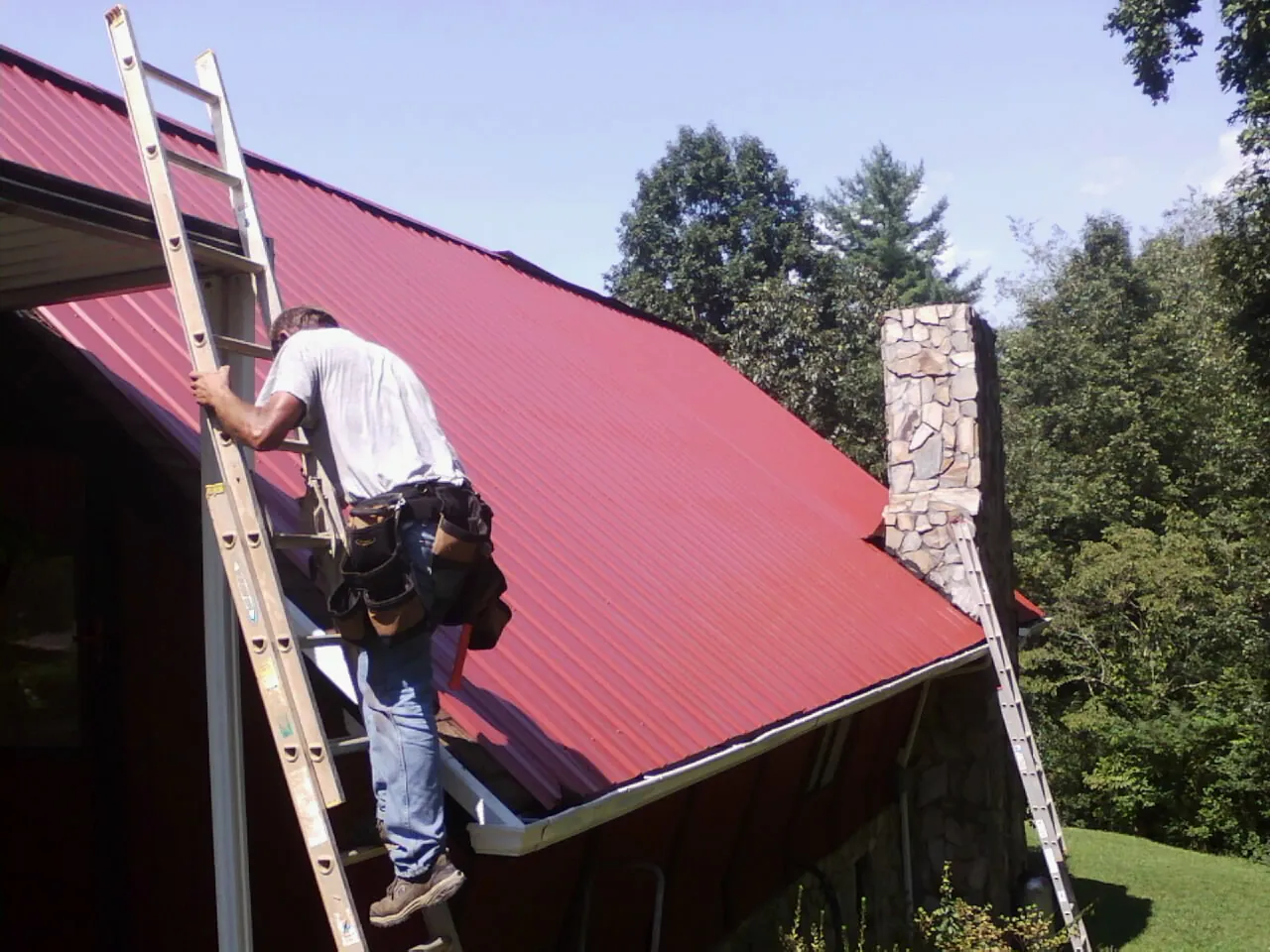 Roofer on a ladder beside a red metal roof and stone chimney — Soffit & Fascia Repair by Roofing Brothers in Fountain Green